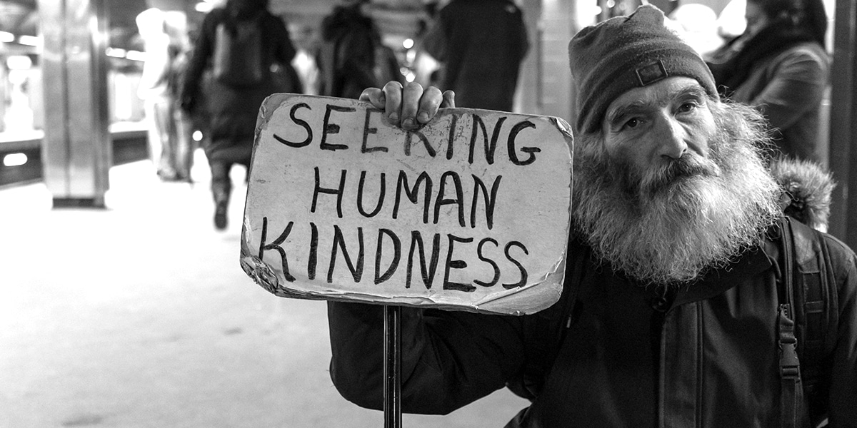 Homeless man holding a sign with "Seeking Human Kindness" written on it.