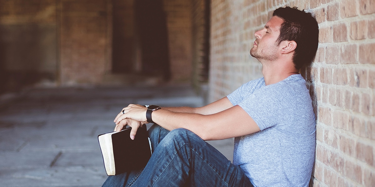 Man praying with his eyes closed while holding a Bible and leaning against a wall