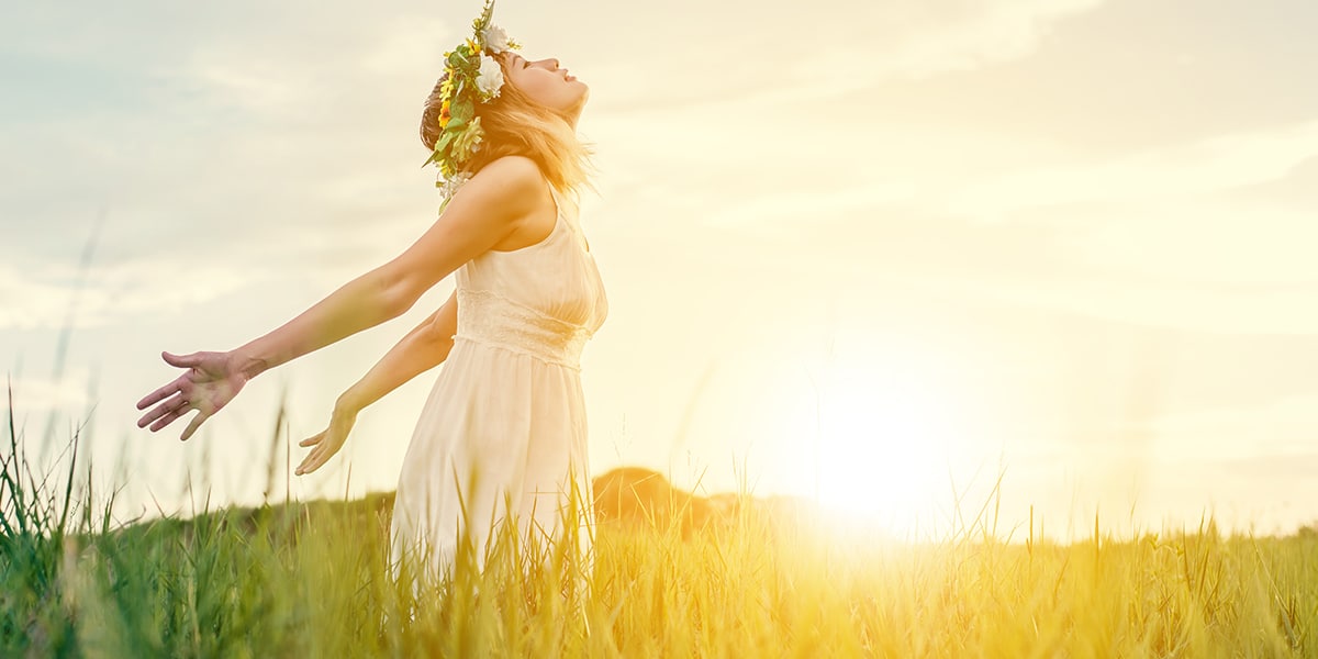 Woman standing in a field during sunset, wearing a flower crown and looking into the sky, reflecting.