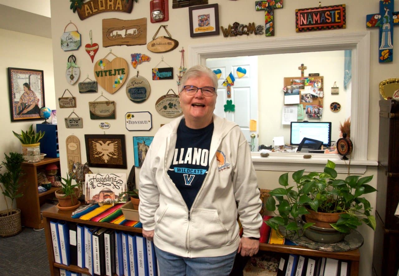 Sister Pat Madden poses in an undated photo before a wall covered with welcome signs given by clients to the Sisters of St. Joseph Welcome Center in Philadelphia. The center offers in-person and online courses in literacy and citizenship and is accredited by the U.S. Department of Justice to assist immigrants with the naturalization process. Some 58 volunteer teachers work with more than 150 students. (OSV News photo/Dan Stockman, Global Sisters Report)