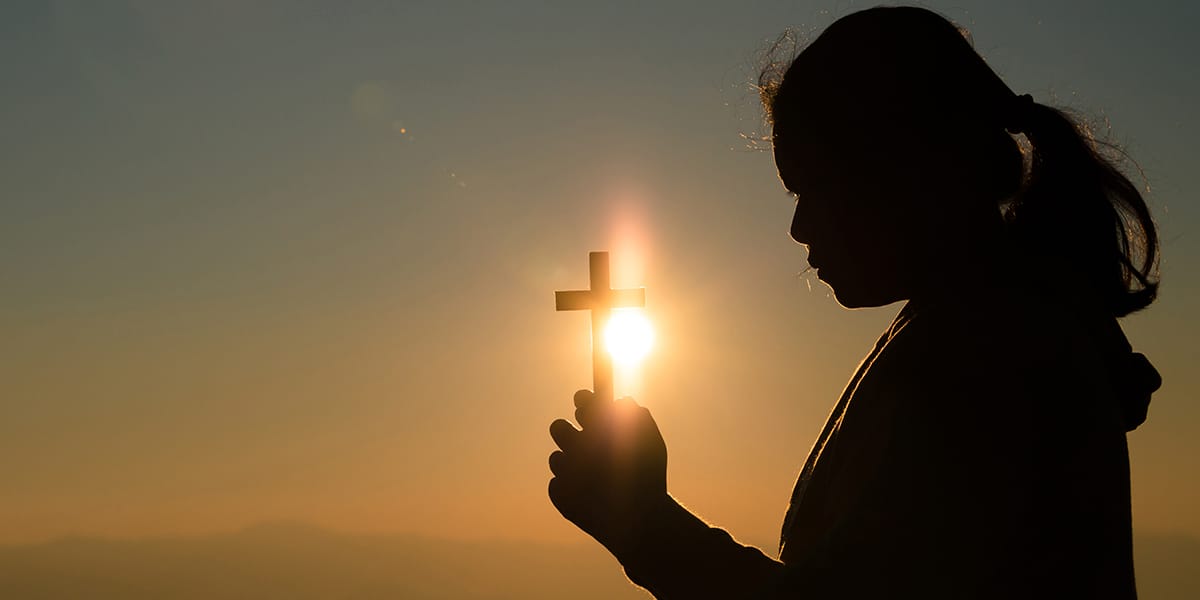 woman holding a cross with the light of a sunset behind the cross