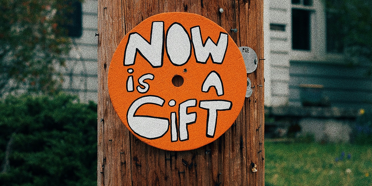 A round sign with the words "Now is a gift" nailed to a wooden pole