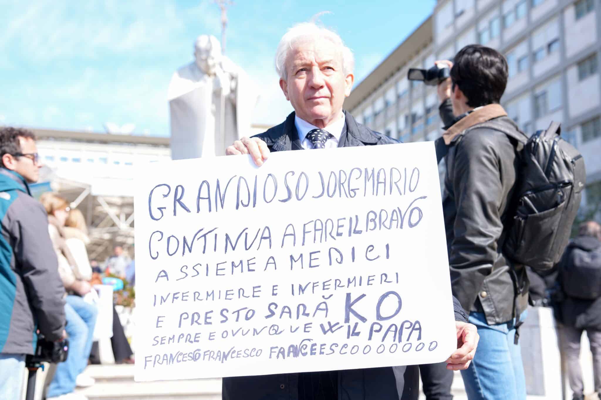 Giuseppe Perazzo holds a sign expressing support for Pope Francis outside Rome’s Gemelli hospital March 2, 2025, while people gather around a statue of St. John Paul II to pray for Pope Francis, who is hospitalized there. (CNS photo/Lola Gomez)