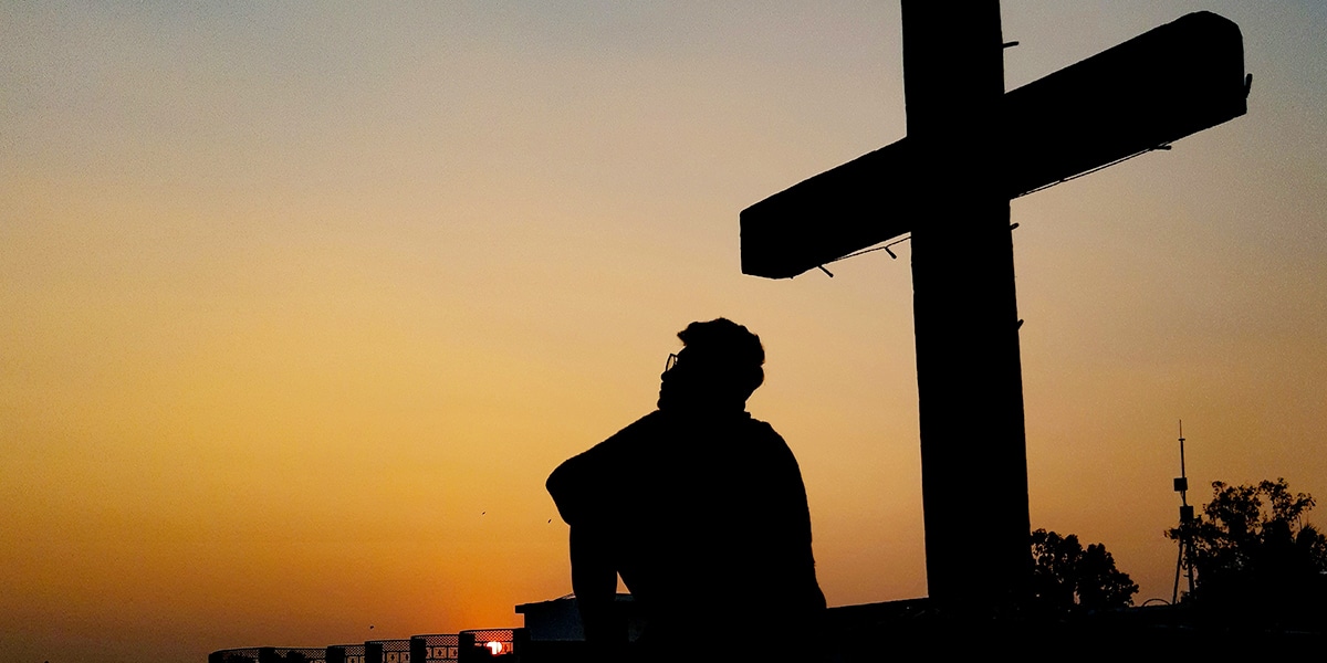 a man sitting in front of a cross with a sunset in the background