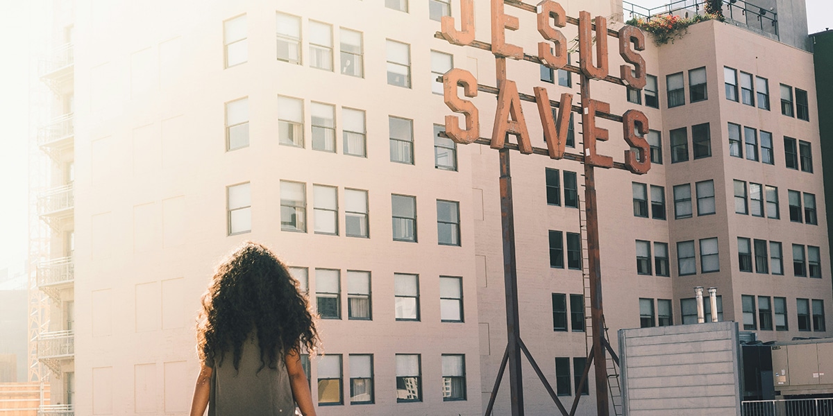 woman looking at a sign "Jesus Saves" surrounded by buildings